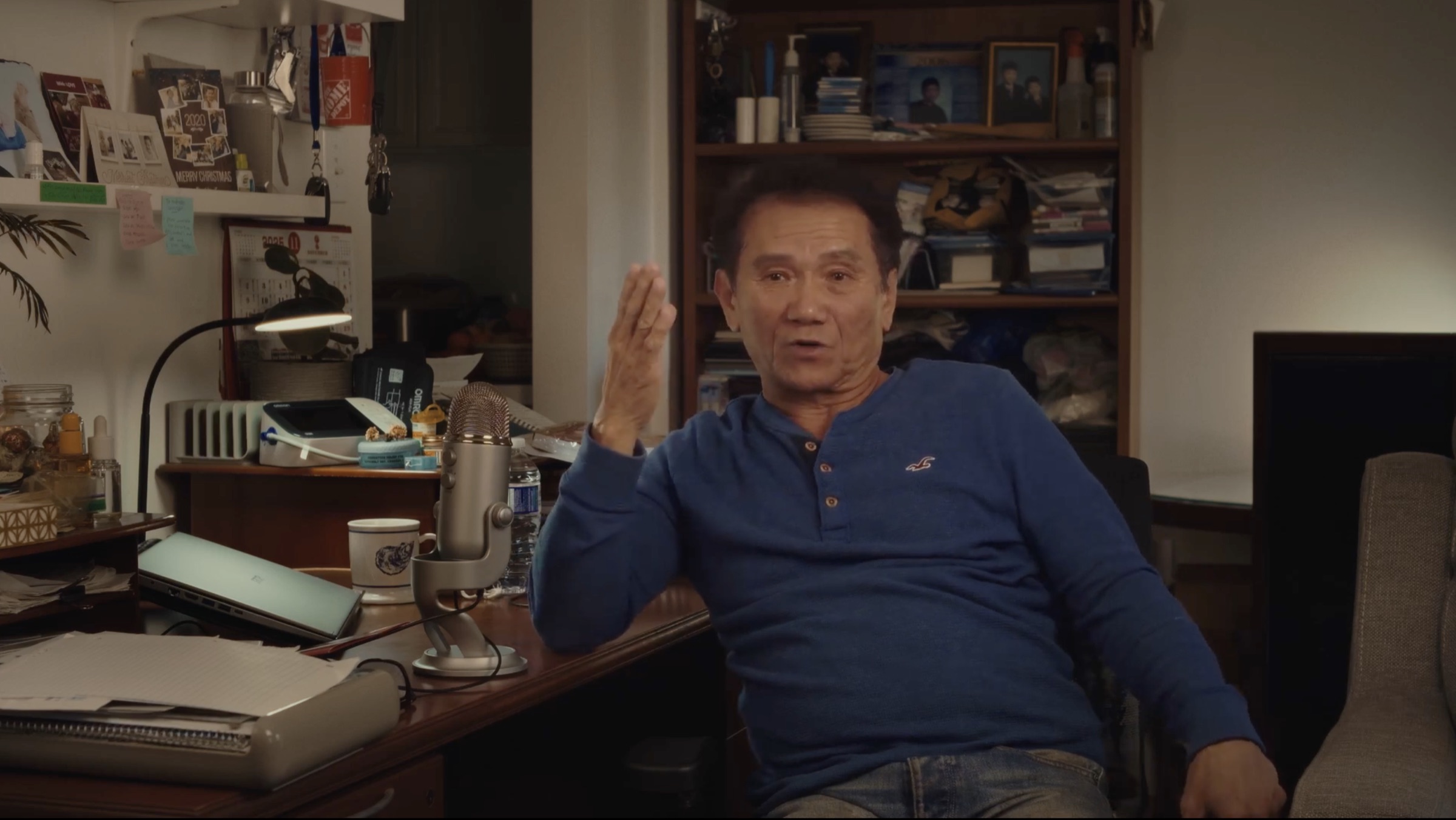 A man at a cluttered home desk mid-sentence, hand raised, a podcast microphone in front of him.