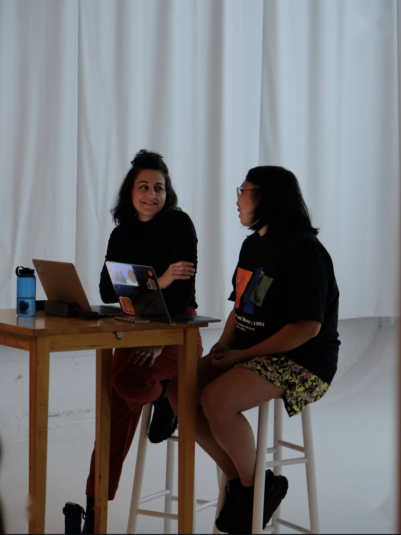 Maya Bankovic and Joyce Wong at a standing desk with laptops in front of a white cyc, mid-conversation.